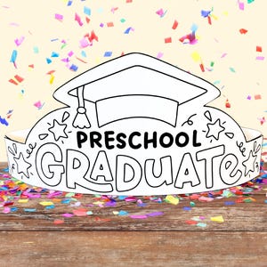 May include: A white paper crown with a graduation cap on top. The crown says "PRESCHOOL GRADUATE" and has stars and other decorations. The crown is sitting on a wooden surface with colorful confetti scattered around it.