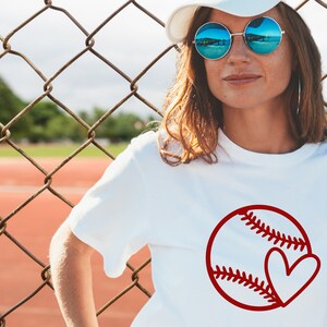 May include: A white t-shirt with a red baseball and heart design. The baseball has white stitching and the heart is inside the baseball.