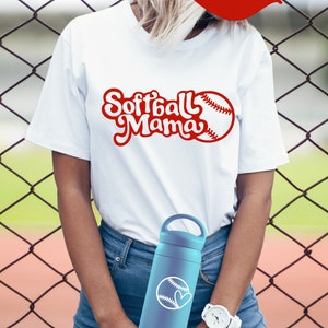 May include: A white t-shirt with the text "Softball Mama" in red cursive lettering and a red baseball graphic. The person is wearing blue jeans and holding a blue water bottle with a white baseball graphic.