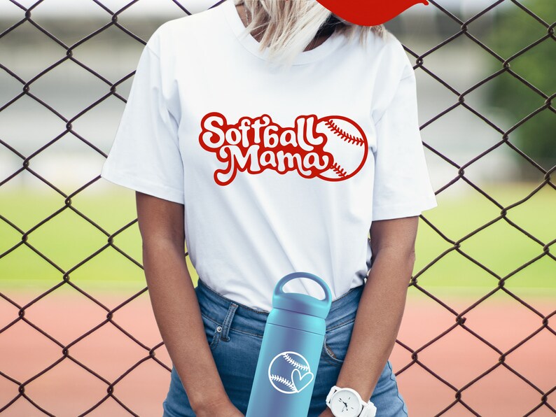 May include: A white t-shirt with the text "Softball Mama" in red cursive lettering and a red baseball graphic. The person is wearing blue jeans and holding a blue water bottle with a white baseball graphic.