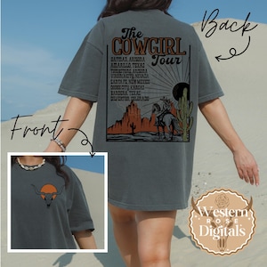 May include: Dark gray t-shirt featuring a bull skull with an orange sun graphic on the front. The back of the shirt displays "The Cowgirl Tour" graphic with a desert scene and tour locations. The shirt is from Western Rose Digitals.