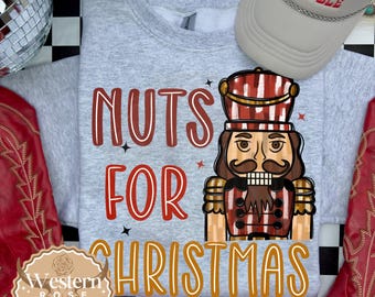 Op de afbeelding: Een grijze sweatshirt met de tekst "NUTS FOR CHRISTMAS" en een cartoon notenkraker afbeelding. De afbeelding bevat ook een beige pet met de tekst "HERE COMES TROUBLE" en rode cowboylaarzen.