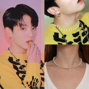 Könnte beinhalten: Nahaufnahme einer Person, die einen gelben Pullover mit schwarzem Muster und eine Perlenkette trägt. Die Person hält eine grüne Traube in der Hand. Das Bild zeigt auch eine Nahaufnahme einer Perlenkette am Hals einer Person.