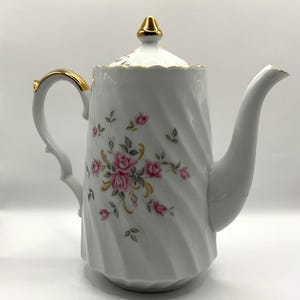 May include: White porcelain coffee pot with a gold trim and a floral design featuring pink roses and green leaves. The pot has a swirling pattern and a lid with a gold knob.