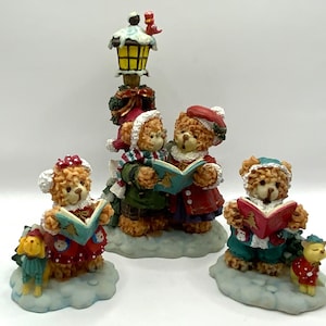 May include: A set of four Christmas figurines featuring bears singing carols under a street lamp. The bears are wearing red, green, and blue clothing and holding songbooks. The street lamp is decorated with a red bird.