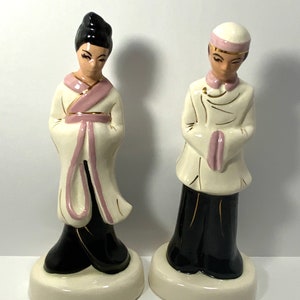 May include: A pair of ceramic figurines depicting a man and woman in traditional Asian clothing. The woman is wearing a white robe with pink accents and a black skirt. The man is wearing a white robe with pink accents and black pants. Both figurines have black hair and are standing on white bases.