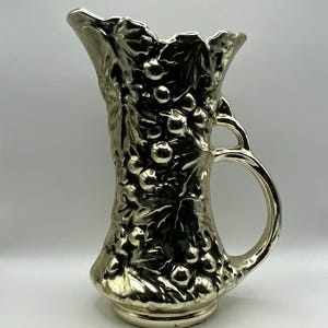 May include: A silver-colored pitcher with a handle, featuring a detailed embossed design of grapes and leaves. The pitcher has a wide opening and a decorative base, suitable for serving beverages or as a decorative item.