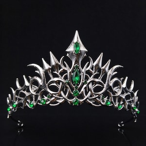Op de afbeelding: Een zilverkleurige metalen tiara met een ingewikkeld, puntig en wervelend ontwerp. Het hoofddeksel is versierd met meerdere kleine, ronde en marquise-geslepen smaragdgroene edelstenen die in het metalen frame zijn gezet.