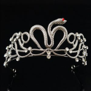 Può includere: Un diadema in metallo color argento con un motivo centrale a forma di serpente e una singola pietra rossa come occhio. La fascia è decorata con intricati motivi a scaglie e numerosi piccoli strass rotondi e trasparenti incastonati.