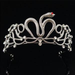 Könnte beinhalten: Ein silberfarbenes Diadem aus Metall mit einem zentralen Schlangenmotiv und einem einzelnen roten Stein als Auge. Das Band ist mit komplexen, strukturierten Schlangenmustern verziert und mit zahlreichen klaren, funkelnden Kristallen besetzt.