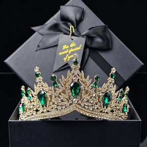 Peut inclure: Une couronne en métal doré ornée de nombreux strass transparents et de grands cristaux vert émeraude en forme de goutte. La couronne est placée devant une boîte cadeau noire avec un ruban noir et une étiquette portant l'inscription For the most special of you.
