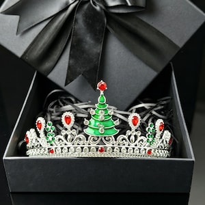 May include: A silver-toned metal tiara featuring a central green enamelled Christmas tree ornament topped with a red crystal. The crown is embellished with clear white rhinestones, red teardrop-shaped crystals, and small green crystal accents.