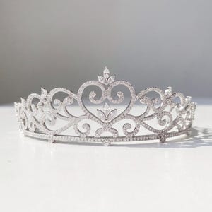 May include: A silver-toned tiara featuring an intricate filigree design with heart-shaped motifs and scrolling patterns. The metal frame is densely encrusted with small clear crystals and accented with larger marquise-cut stones.