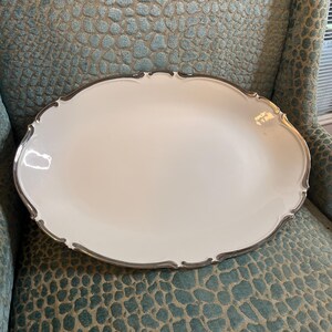 May include: A white, oval serving platter with a silver-coloured, scalloped edge. The platter is set against a textured, teal-coloured background, suggesting a vintage or antique aesthetic. The design is simple and elegant.