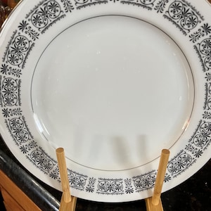 May include: A white ceramic plate with a black and white geometric pattern around the edge. The plate is displayed on a wooden stand.