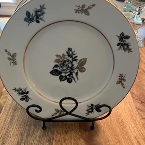 Könnte beinhalten: Ein Set aus sechs weißen Speisetellern mit goldfarbenem Rand und schwarzem und goldenem Blumendesign. Die Teller sind auf einem schwarzen Metallständer gestapelt.