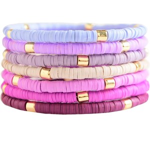 Op de afbeelding: Een set van acht kleurrijke armbanden gemaakt van kleine, ronde kralen. De armbanden zijn op elkaar gestapeld, met kleuren die overgaan van lichtblauw naar paars. Elke armband heeft een gouden kraal die de gekleurde kralen scheidt.