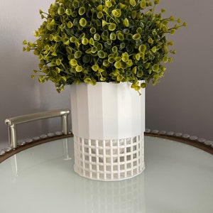 Puede incluir: Maceta blanca con un patrón de rejilla, que contiene un arbusto artificial verde con pequeñas hojas redondas.