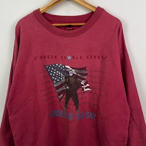 Può includere: Una felpa bordeaux con la scritta "FADED GLORY" e una grafica di una persona che tiene una bandiera americana. La frase "HEROES OF THE SKY" è stampata sotto. La felpa è appesa a una gruccia di legno.