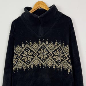 May include: Black fleece pullover with a high collar and a quarter-zip closure. The sweater features a geometric pattern in tan across the chest. The Timberland logo is visible on the lower right side. The garment is hanging on a wooden hanger.
