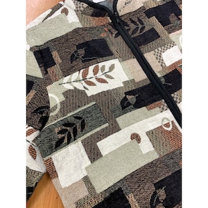 Può includere: Una giacca con motivo marrone, nero e crema con chiusura a zip. La giacca ha un design geometrico con foglie e forme astratte.