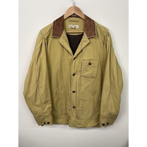 Puede incluir: Chaqueta color beige con cuello de cuero marrón, cierre de botones y dos bolsillos delanteros. La chaqueta está colgada de una percha de madera sobre un fondo blanco. La chaqueta tiene un diseño clásico y utilitario.