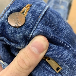 May include: Close-up of a blue denim jean with a copper button and a gold zipper pull that says "SCOVILL".