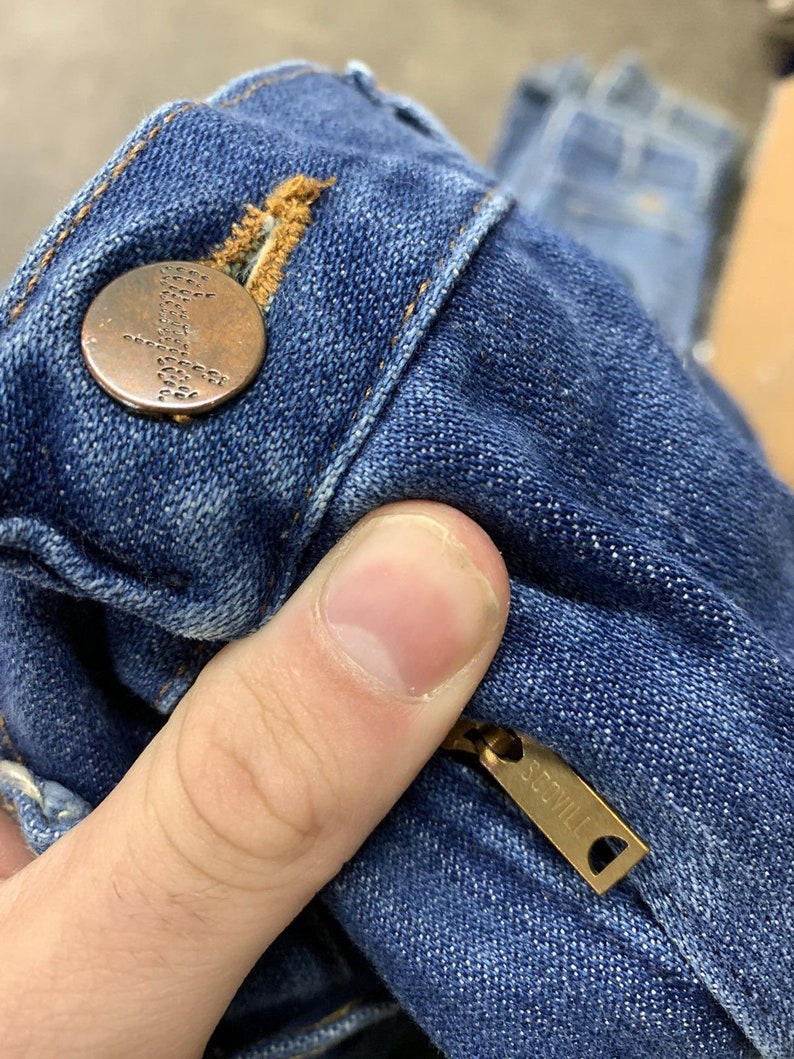 May include: Close-up of a blue denim jean with a copper button and a gold zipper pull that says "SCOVILL".