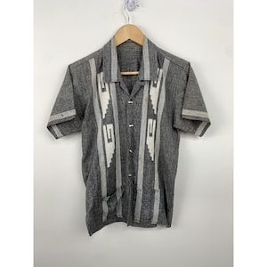 May include: A gray short-sleeved button-up shirt with white geometric embroidery. The shirt has a collar and a pocket on the left side.