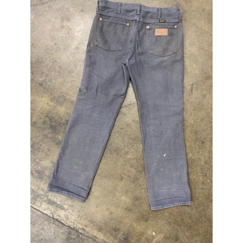 May include: A pair of dark gray denim jeans with a brown leather patch on the back right pocket. The patch has the word "Wrangler" printed on it.