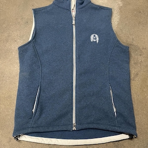 May include: A blue fleece vest with a full-length zipper and two side pockets. The vest has a white trim along the collar, armholes, and hem. A small emblem is on the upper left chest.