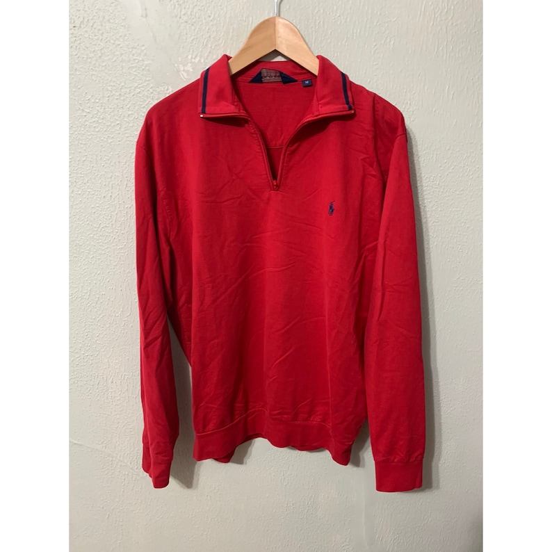 May include: A red long-sleeve sweater with a blue polo player logo and a half-zip closure. The sweater has a navy blue trim around the collar.