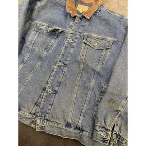 Op de afbeelding: Een blauwe denimjas met een bruine corduroykraag. De jas heeft een knoopsluiting en twee voorzakken.
