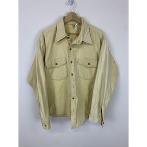 Pode incluir: Uma camisa de botão de manga comprida de cor amarelo claro com dois bolsos no peito e um fecho de botão único no colarinho.
