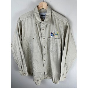 May include: A beige long-sleeve button-up shirt with a pocket on the left chest. The pocket has an embroidered logo of a blue cartoon character with the text "CURRY'S 1979 MUSIC COLLECTION".