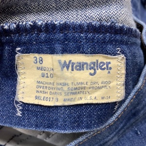 May include: A close-up of a blue denim Wrangler jeans label with white stitching. The label has the text "38", "910", "Wrangler", "MACHINE WASH, TUMBLE DRY, AVOID OVERDRYING REMOVE PROMPTLY. WASH DARKS SEPARATELY.", "9KLE0173 MADE IN U.S.A. W-14".