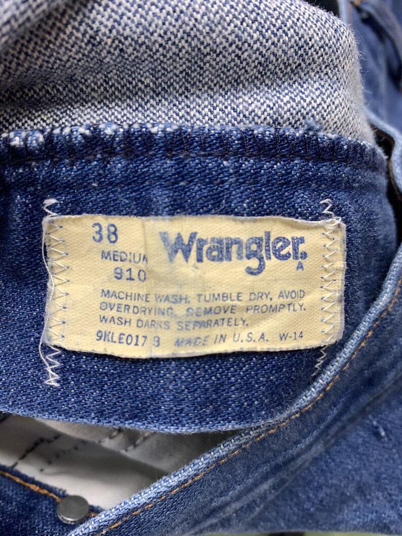 May include: A close-up of a blue denim Wrangler jeans label with white stitching. The label has the text "38", "910", "Wrangler", "MACHINE WASH, TUMBLE DRY, AVOID OVERDRYING REMOVE PROMPTLY. WASH DARKS SEPARATELY.", "9KLE0173 MADE IN U.S.A. W-14".