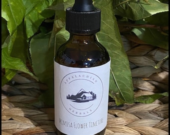May include: A brown glass bottle with a black dropper top and a white label that reads "Appalachian Herbals Mimosa Flower Tincture". The bottle is sitting on a woven wicker surface.