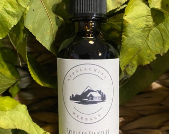 May include: A glass bottle with a black dropper top and a white label. The label says "Appalachian Herbals Skullcap Tincture". The bottle is surrounded by green leaves.