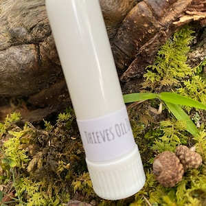 May include: A white roll-on bottle with a white label that says "Thieves Oil" on it. The bottle is sitting on a bed of green moss.