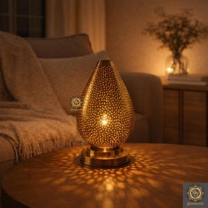 Può includere: Una lampada da tavolo dorata a forma di goccia con un design traforato. La lampada poggia su una base rotonda e proietta una luce a motivi sul tavolo. La lampada è accesa, creando un'atmosfera calda.