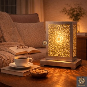 Può includere: Lampada decorativa in metallo argentato con un motivo geometrico, che emette una calda luce gialla. La lampada è su un tavolo di legno, accanto a una tazza di caffè, un libro e una ciotola di noci. Il motivo è intricato.