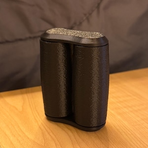 Peut inclure: Un récipient noir à deux chambres avec un dessus texturé et moucheté. Le récipient est cylindrique avec une base arrondie et repose sur une surface en bois clair. L'arrière-plan est un tissu sombre et texturé.