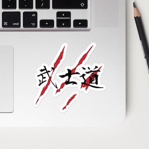 May include: A silver laptop with a sticker featuring the Japanese kanji for Bushido in black ink, layered over three jagged red claw marks. The laptop is positioned next to a black pencil, a blue ballpoint pen, a smartphone, and a red square note.