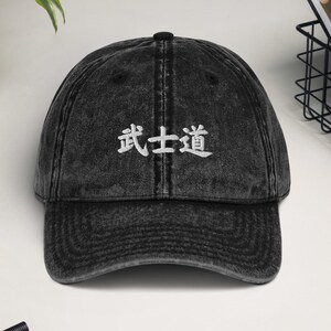 May include: A black acid-wash baseball cap featuring white embroidered Japanese kanji characters that read Bushido on the front panel. The cap has a curved brim, a button on top, and stitched eyelets for ventilation, presenting a worn-in texture.