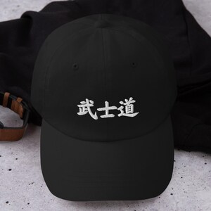 May include: Black baseball cap with the white Japanese characters for Bushido embroidered on the front. The cap is made of a soft material and has a curved brim. A watch with a brown leather band is visible in the background.