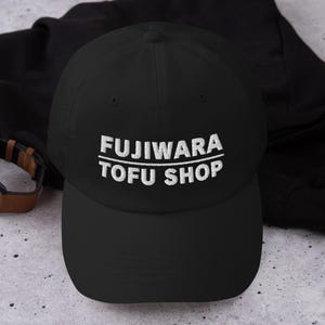Op de afbeelding: Een zwarte baseballpet met de wit geborduurde tekst FUJIWARA TOFU SHOP op het voorpaneel. De pet heeft een gestructureerde kroon, een gebogen klep en een knoop aan de bovenkant. Een horizontale lijn scheidt de twee regels tekst van elkaar.