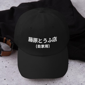 Op de afbeelding: Een zwarte honkbalpet met wit geborduurde Japanse tekst die Fujiwara Tofu Shop op de bovenste regel en Privégebruik op de onderste regel leest. De pet heeft een gestructureerde kroon, een gebogen klep en een knoopje aan de bovenzijde.