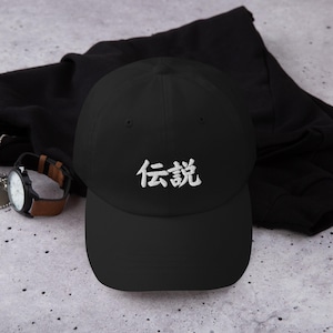 May include: A black baseball cap featuring white embroidered Japanese kanji characters that read 伝説 on the front panel. The cap has a structured crown, a curved brim, and a button on top, presented with a black garment and a wristwatch.
