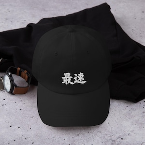 May include: A black baseball cap featuring white embroidered Japanese kanji characters that read 最速 on the front panel. The cap has a structured crown, a curved brim, and a button on top, showcasing a clean and simple design for everyday headwear.